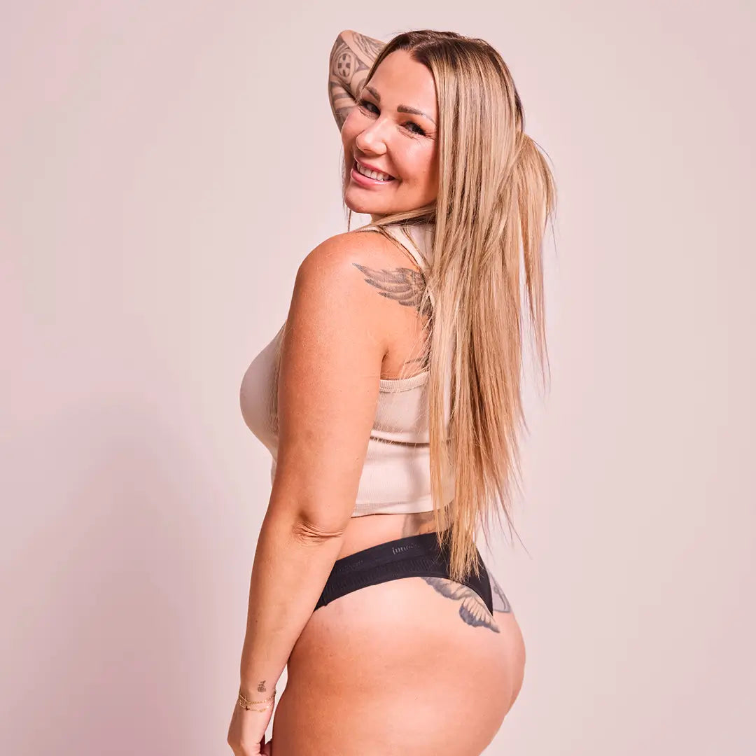 Blonde woman with tattoos in a beige top and the Ribbed Cotton String, smiling over her shoulder