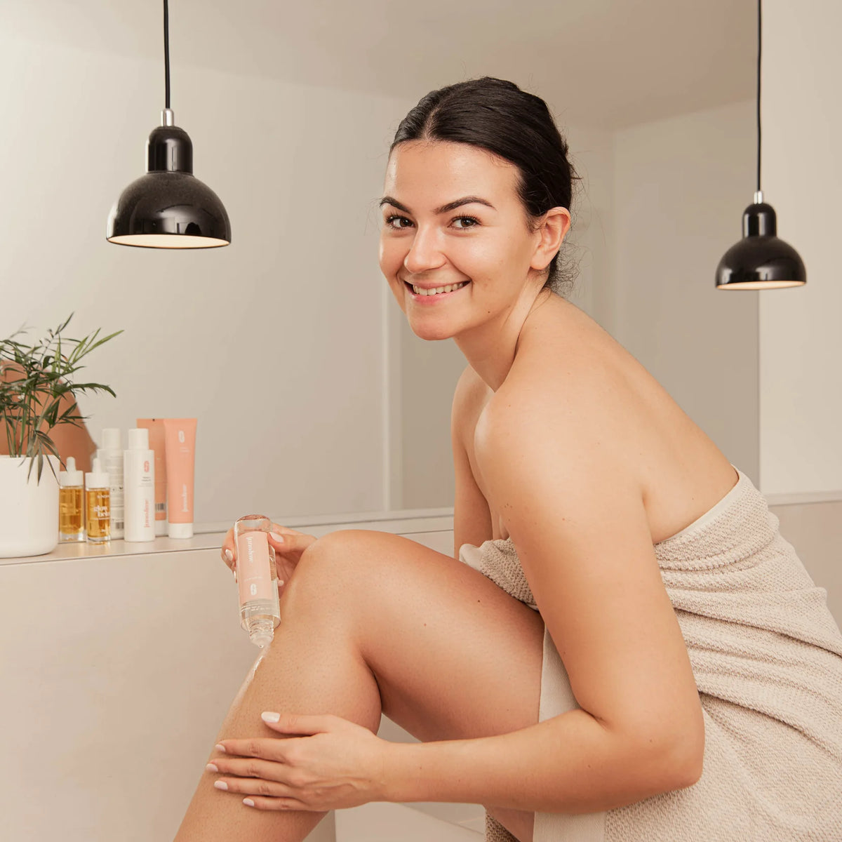 Lächelnde Frau im Handtuch pflegt ihre Beine mit Körperöl im Badezimmer, Wochenbett-Checkliste
