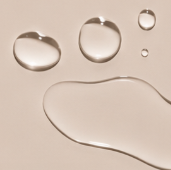 Klare Tropfen des Dry Touch Body Oil aus dem Touch Me Duo auf beigem Grund, juno&me