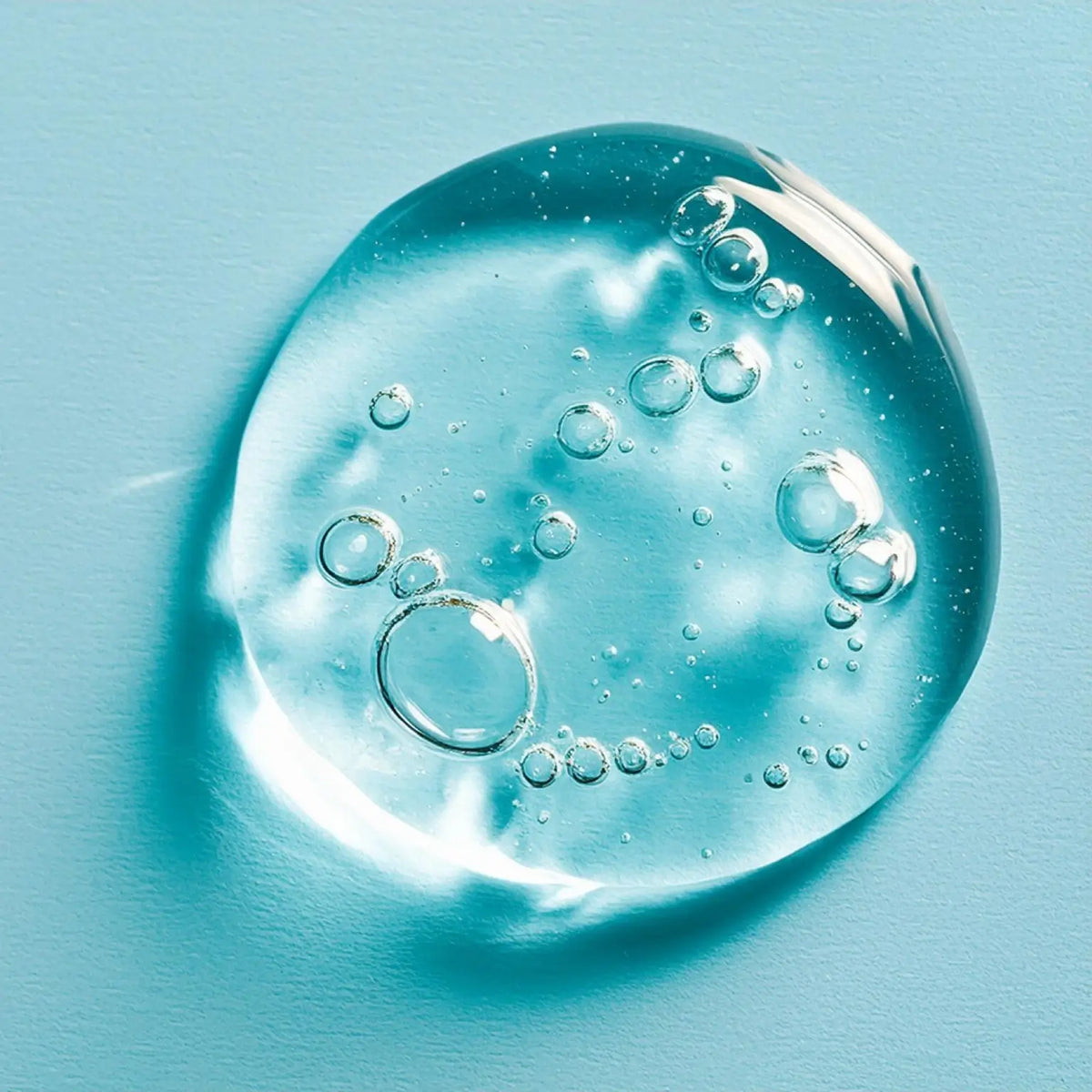 Klarer, gelartiger Tropfen mit Luftblasen ruht auf hellem, blauen Hintergrund.

