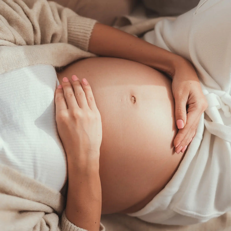 Schwangere Frau hält sanft ihren Bauch, beige Kleidung.
