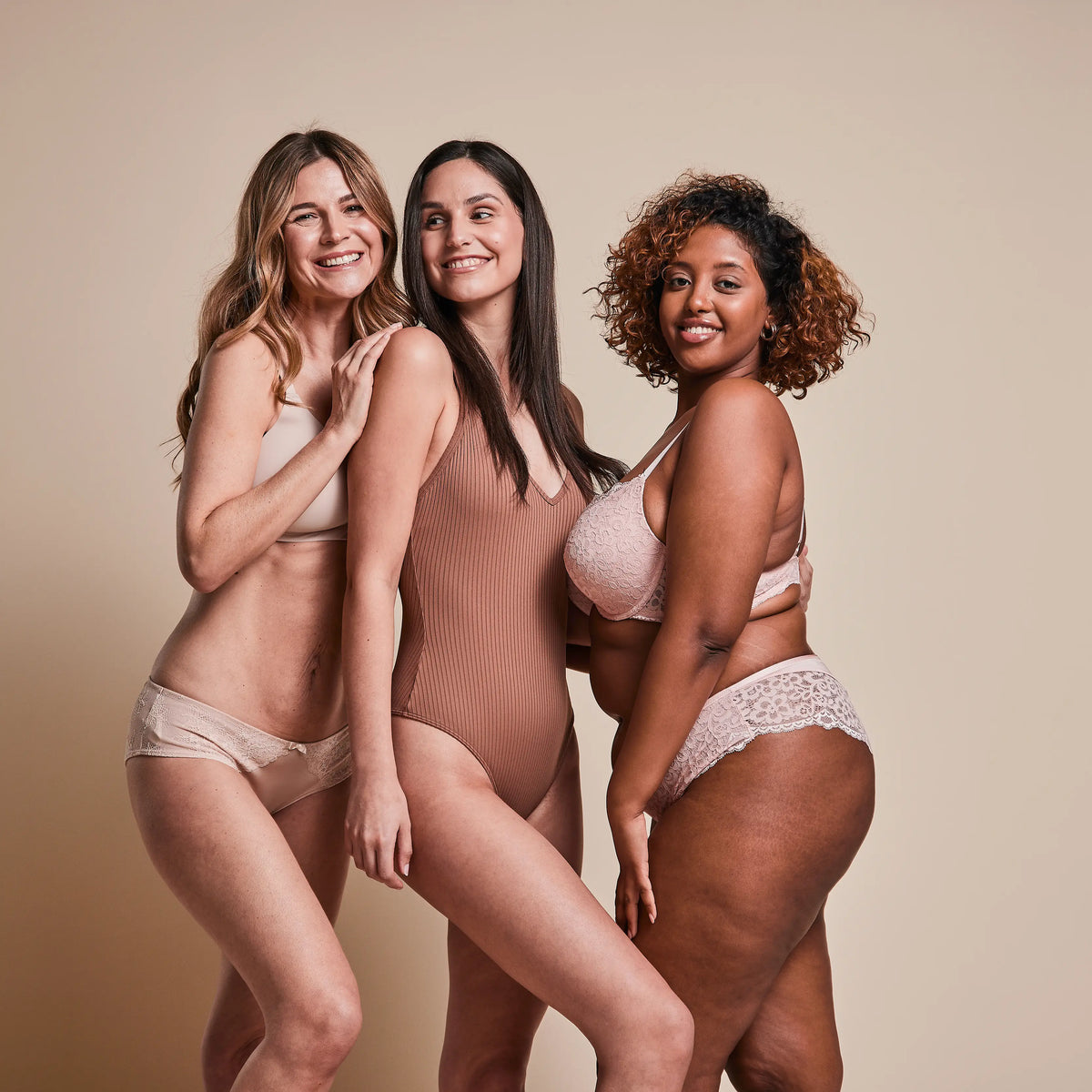 Three women in underwear smiling and posing against a beige background.